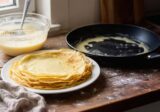 Stos cienkich naleśników na talerzu obok patelni i miski z ciastem naleśnikowym